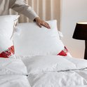 Twee witte kussens met bruine biezen op een bed. Een hand schikt de kussens. Een rode "Canada Montreal" label is zichtbaar.