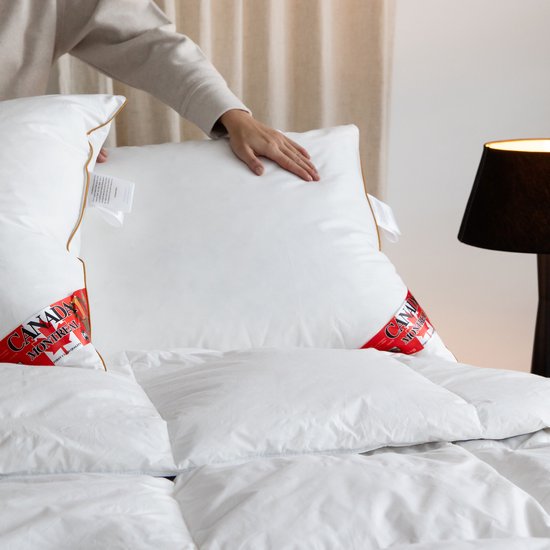 Twee witte kussens met bruine biezen op een bed. Een hand schikt de kussens. Een rode "Canada Montreal" label is zichtbaar.