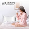 Vrouw in roze badjas drinkt koffie op een bed met witte satijnen kussenslopen. Tekst: Luxe en stijlvol.