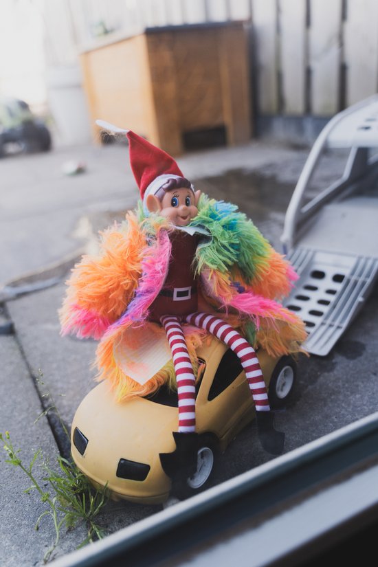 Elf met kerstmuts, rood shirt, gestreepte broek en kleurrijke bontjas, zittend op een gele speelgoedauto.