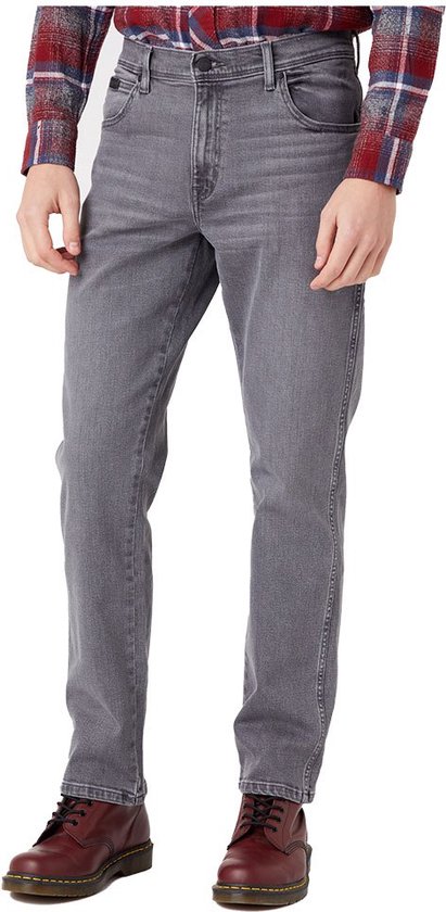 Man staat vol in beeld in een grijze jeans en een rood, blauw geruite blouse. Hij draagt donkerbruine schoenen.