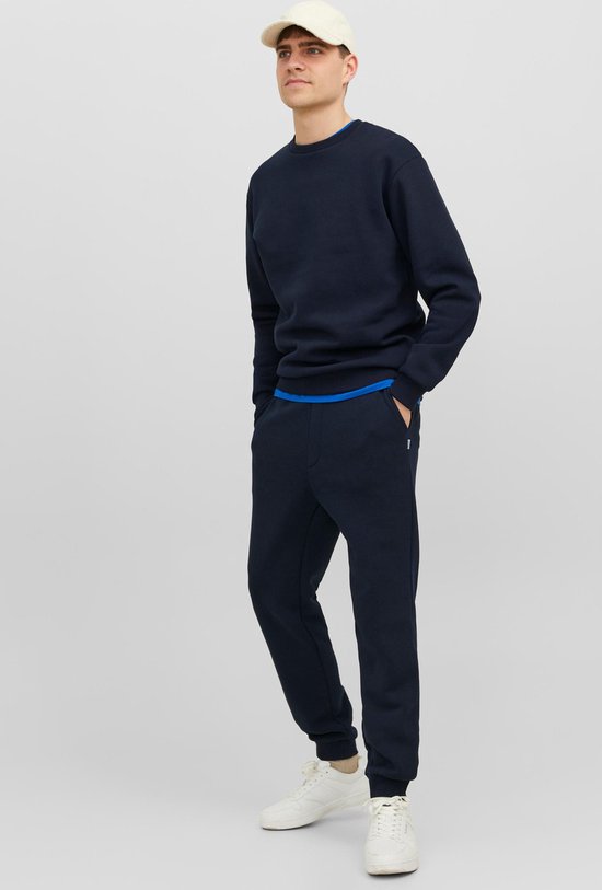 Man in een donkerblauwe sweater en joggingbroek, met een beige pet en witte sneakers.