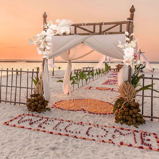 Strandceremonie met houten boog versierd met witte bloemen, roze rozen, pampasgras en witte stof, met 'Welcome' in rozenblaadjes.