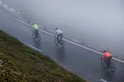Drie fietsers rijden door mist op een bergweg. Eén in een witte jas, één in neongeel en één in oranje.