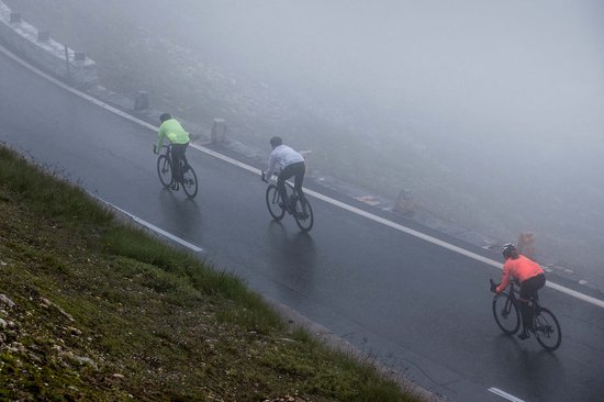 Drie fietsers rijden door mist op een bergweg. Eén in een witte jas, één in neongeel en één in oranje.