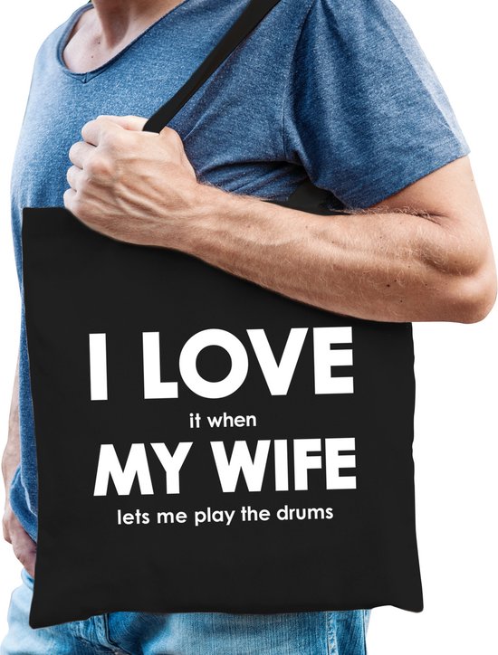 Sac fourre-tout noir en coton avec le texte : 'I LOVE it when MY WIFE lets me play the drums'. Porté par un homme.