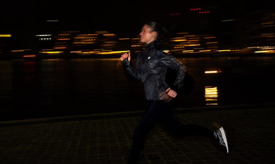Vrouw in zwart hardloopjack met reflecterende, gestreepte print, rent 's nachts langs het water.