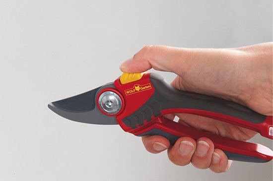 Hand met snoeischaar: rood-grijze soft-inleg handgrepen, gele ontgrendelknop, metalen snijblad.