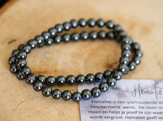 Twee zwarte, glanzende hematiet kralenarmbanden op een houten ondergrond met een kaartje met tekst.