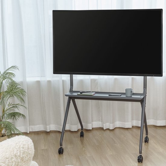 Support TV gris avec un grand écran noir, une étagère multimédia, sur un plancher en bois avec des rideaux blancs.