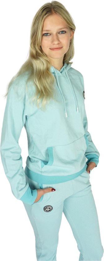 Model in een lichtblauwe joggingbroek en hoodie met capuchon, met opgestreken logo op de broek.