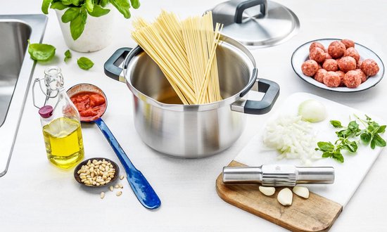 RVS kookpot met deksel, omringd door ingrediënten voor pasta: spaghetti, gehaktballetjes, tomatensaus, olie, ui, knoflook en kruiden.