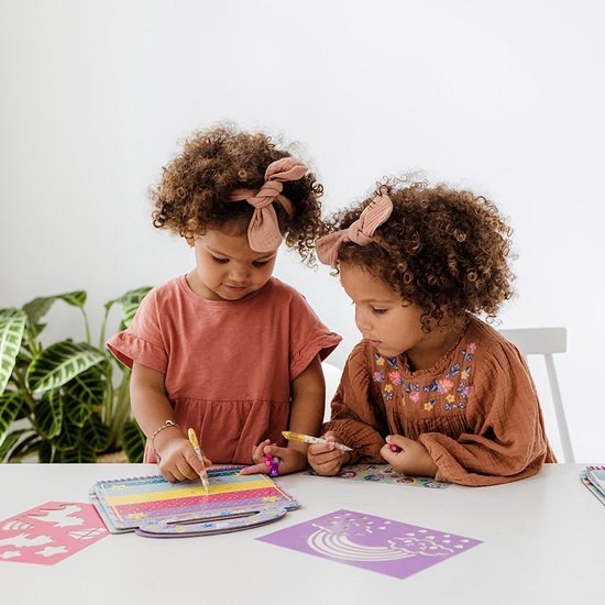 Deux jeunes filles avec des bandeaux et des planches à repasser dessinent dans un livre d'activités Mini MyMy sur le thème de l'arc-en-ciel.