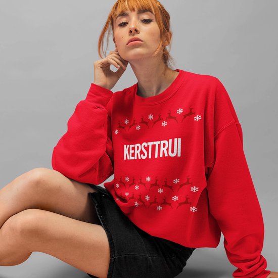 Une jeune femme porte un pull de Noël rouge avec le mot 'KERSTTRUI' et un motif de rennes et de flocons de neige.