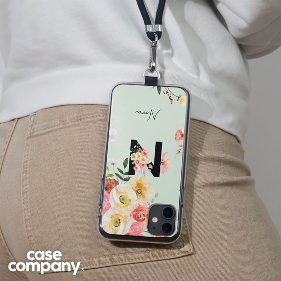 Een persoon draagt een lichtgroen telefoonhoesje met een 'N', bloemen en de naam 'Nora' aan een zwart koord.