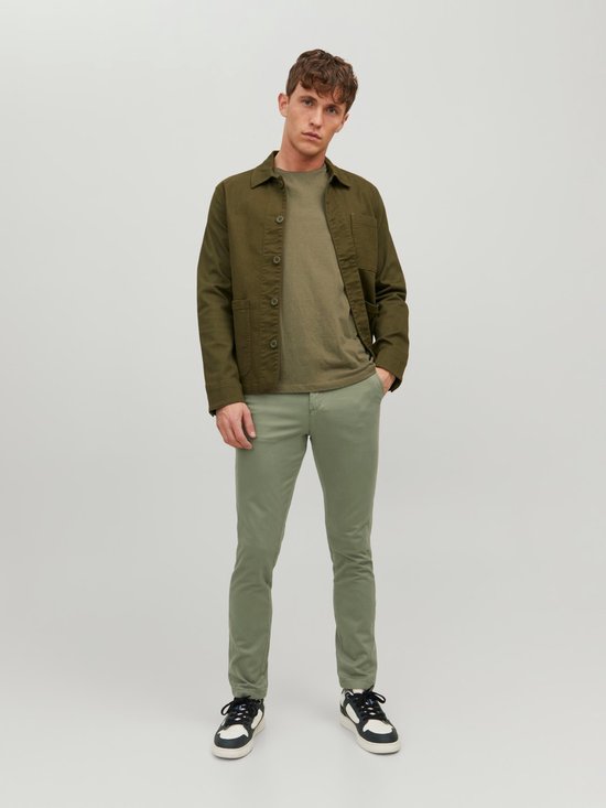Model staand, draagt een olijfgroene chino broek met groen jack en t-shirt.