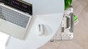 Witte Logitech MX Anywhere 3S muis op een wit bureau naast een laptop en een zilveren koffer.