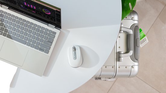 Witte Logitech MX Anywhere 3S muis op een wit bureau naast een laptop en een zilveren koffer.