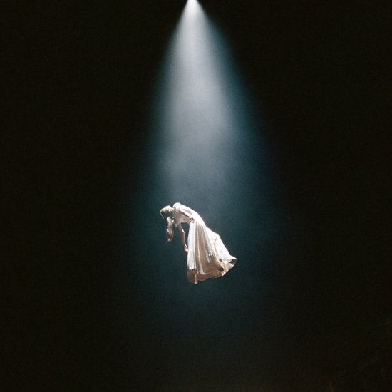Een vrouw in een witte jurk zweeft in een donkere ruimte, verlicht door een enkele lichtstraal van boven.