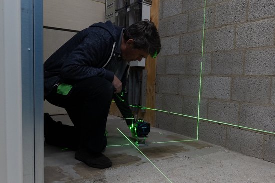 Man met ProXtar kruislijnlaser op een statief, projecteert groene laserlijnen op een betonnen muur en vloer.
