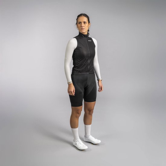 Vrouw met lange donkere haren in paardenstaart, draagt zwarte mouwloze fietsjas, lange witte mouwen, zwarte fietsbroek en witte sokken.