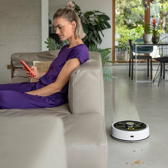 Witte robotstofzuiger met zwarte bovenkant, met kringen op de vloer. Een vrouw zit op een bank met een telefoon.