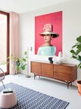 Audrey Hepburn portant un grand chapeau, un nœud rose et un haut pâle, sur fond rose, près d'un buffet en bois et d'une fenêtre.