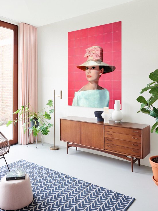 Audrey Hepburn portant un grand chapeau, un nœud rose et un haut pâle, sur fond rose, près d'un buffet en bois et d'une fenêtre.