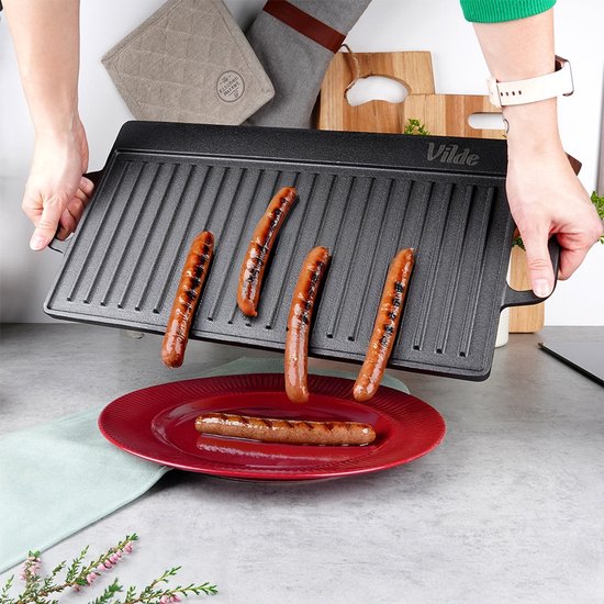 Gegrilde worstjes op een zwarte gietijzeren grillplaat die boven een rode schaal wordt gehouden.