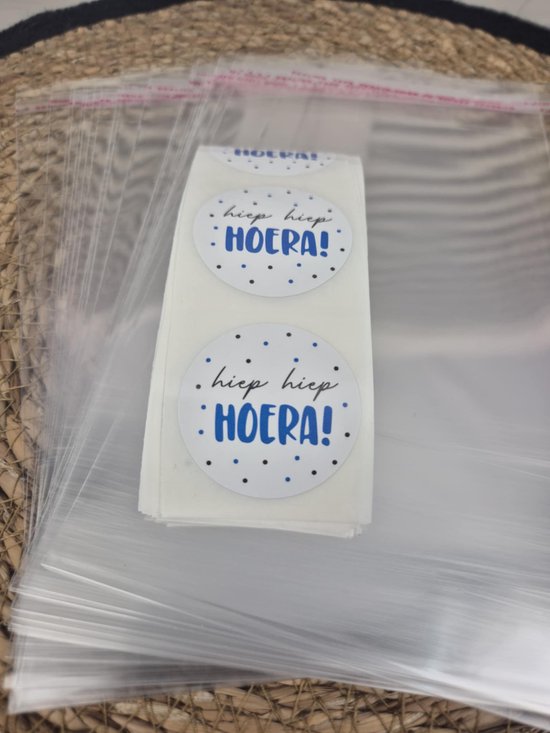 Rol witte stickers met blauwe 'hiep hiep HOERA!' tekst en blauwe stipjes, naast transparante celofaan zakjes met rode plakrand.