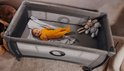 Een baby in een oranje outfit ligt in een grijs campingbedje met sterren, samen met twee knuffels.