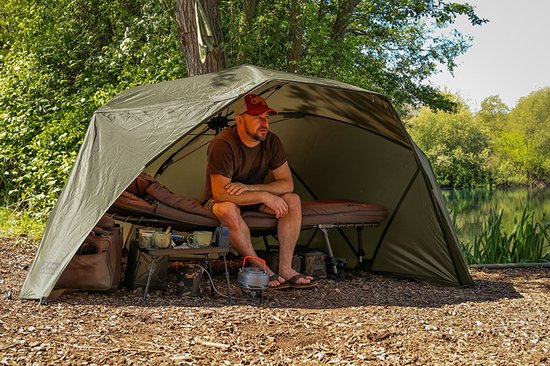 Man in olijfgroene visbrolly, zittend op een bedchair met een waterkoker en mokken op een tafeltje.