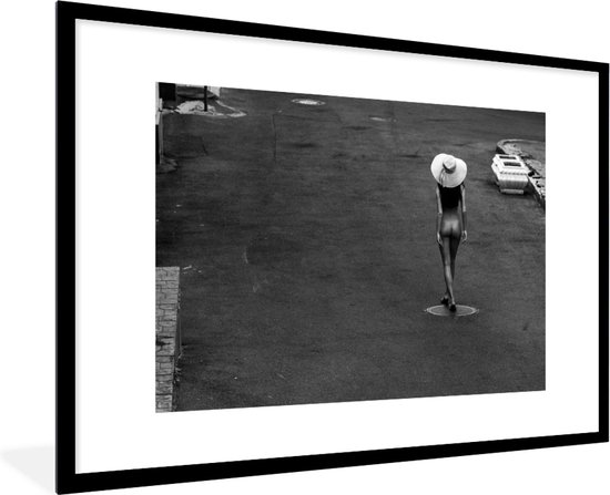 Photo noir et blanc d'une femme portant un chapeau de paille, pieds nus dans la rue, avec des bateaux en arrière-plan.