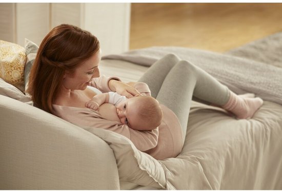 Vrouw met bruin haar voedt een baby op een bed. Ze draagt een roze top en grijze legging.