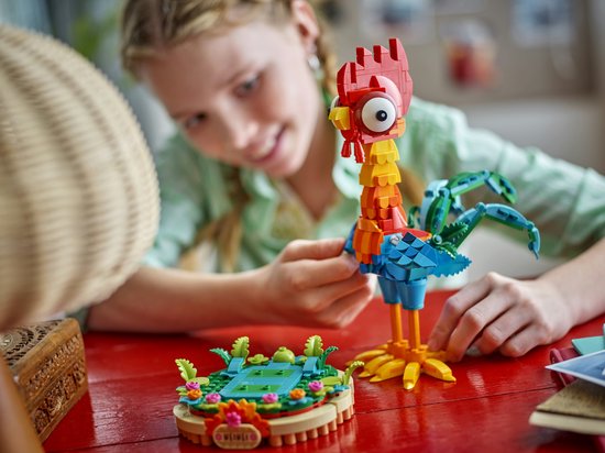 Une fille construit la figurine Heihei avec des LEGO, avec une tête rouge et des ailes et une queue bleu-vert.
