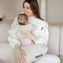 Vrouw met blonde haren, draagt een witte coltrui en houdt een baby vast op een beige voedingskussen.
