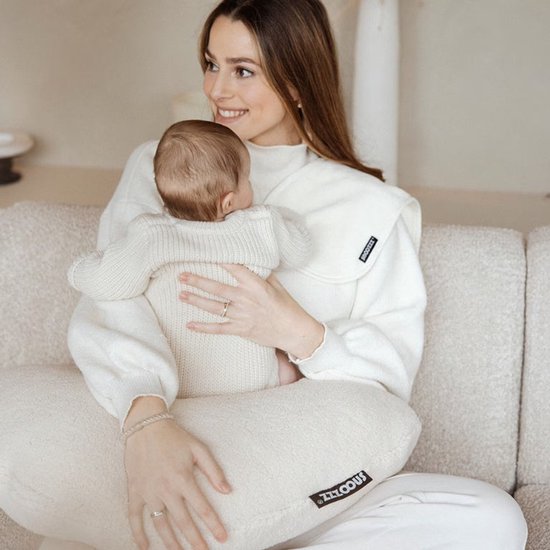 Vrouw met blonde haren, draagt een witte coltrui en houdt een baby vast op een beige voedingskussen.