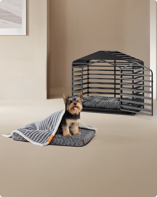Yorkshire Terriër zit op een donkergrijs pluche hondenbed met geborduurde bloemmotieven, bedekt met een geribbelde grijze deken, naast een hondenkennel.