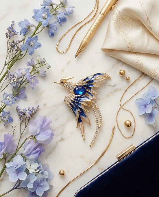 Gouden broche in vogelvorm met blauwe steen en kristallen, omringd door blauwe en paarse bloemen en gouden accessoires.