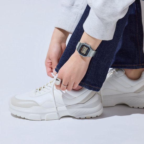 Close-up van handen van een persoon die een spijkerbroek en witte sneakers draagt, met een zilverkleurig Casio horloge om de linkerpols.