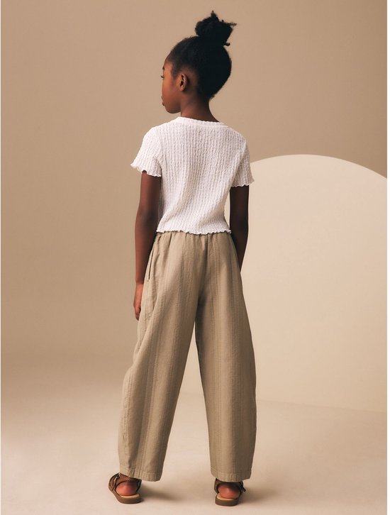 Meisje met bruine huid in een witte top met korte mouwen en een beige broek met verticale strepen.