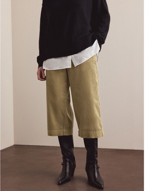 Model in een bruine corduroy broek en zwarte trui, met zwarte laarzen.