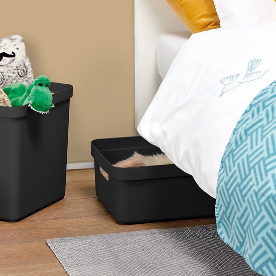 Twee zwarte opbergboxen, één hoog met groene dinosaurus, één laag gevuld met vachtjes, naast een bed met wit beddengoed.