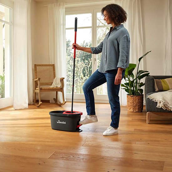 Vrouw in blauwe blouse en jeans gebruikt de rode mop met pedaal in een woonkamer.
