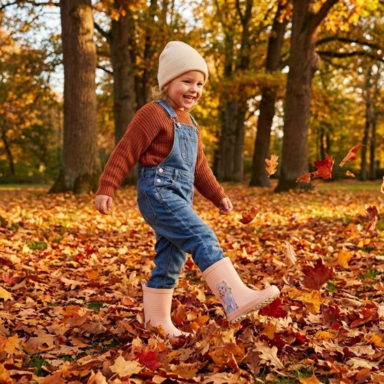 Een lachend meisje in een herfstbos, spelend in de bladeren met lichtroze Frozen regenlaarzen aan.