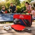 Een gezin speelt tafeltennis in de tuin. Op de voorgrond liggen twee rode batjes en drie witte ballen naast een rode draagtas met Slazenger logo.