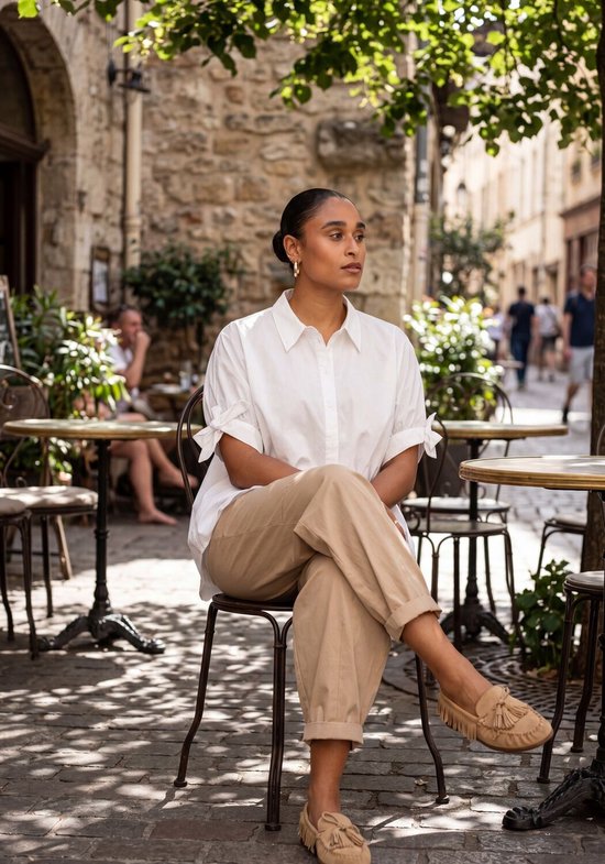 Vrouw in beige broek en witte blouse zit buiten op een stoel.