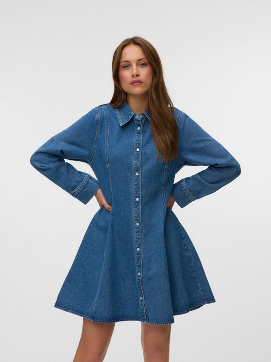Model draagt een blauwe denim jurk met lange mouwen en een uitlopende rok. Ze heeft haar handen in haar zij.