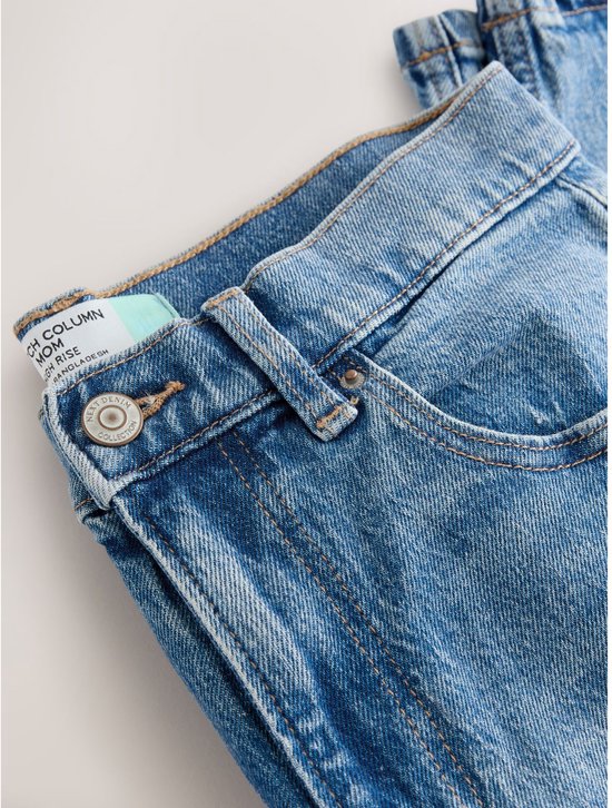Detailopname van een spijkerbroek met hoge taille, met een knoopsluiting en een label met 'COLUMN MOM HIGH RISE'.