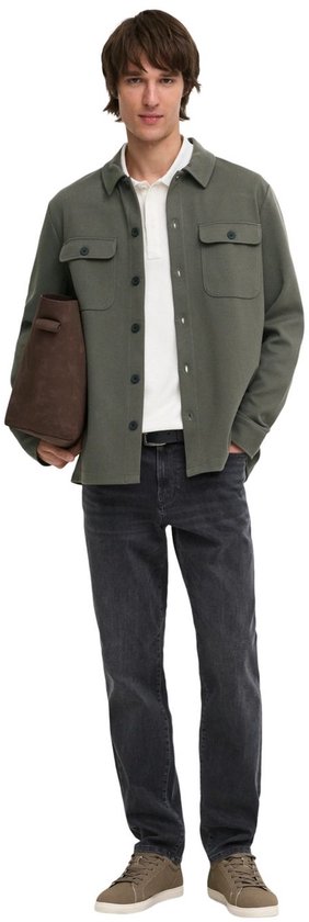 Man in olijfgroene utility blouse, wit poloshirt, grijze jeans, bruine tas en sneakers.